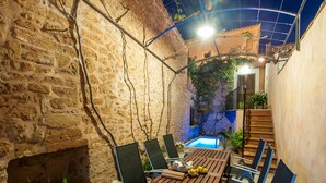 Une terrasse extérieure aux murs de pierre, avec une table et des chaises en bois, une petite piscine et un escalier menant à un niveau supérieur.