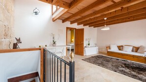 Une pièce spacieuse avec un plafond haut, des poutres en bois, un mur en pierre, un canapé blanc, une petite table et un escalier doté d’une rampe.