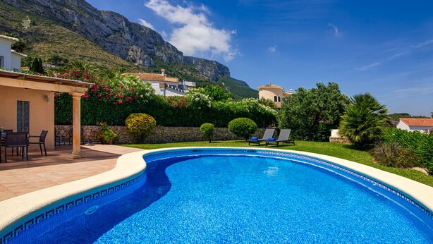 Una piscina con borde de azulejos, rodeada por un patio con sillas y una mesa, y una vista de una montaña al fondo.