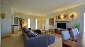 Una sala de estar moderna con un sofá, una mesa de comedor y un televisor. Se observa un área de cocina al fondo.