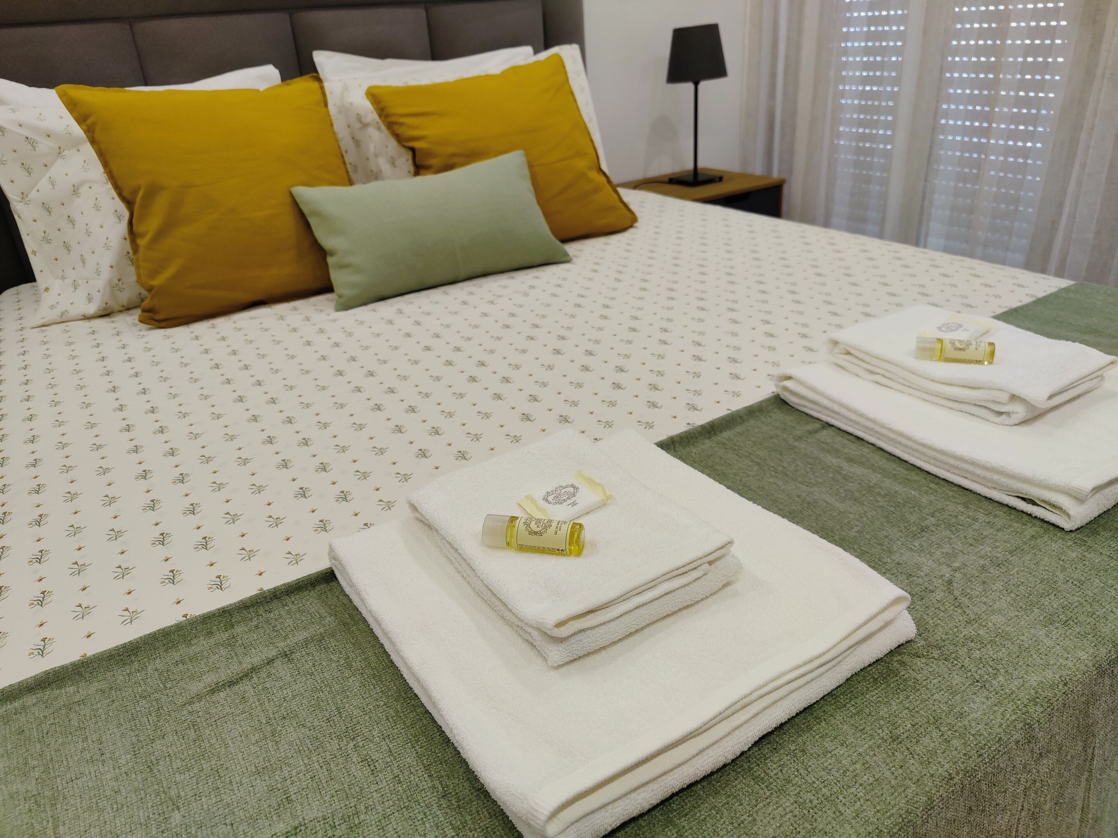 Una cama bien hecha con una colcha estampada, dos almohadas y toallas dobladas, junto con una botella de loción.