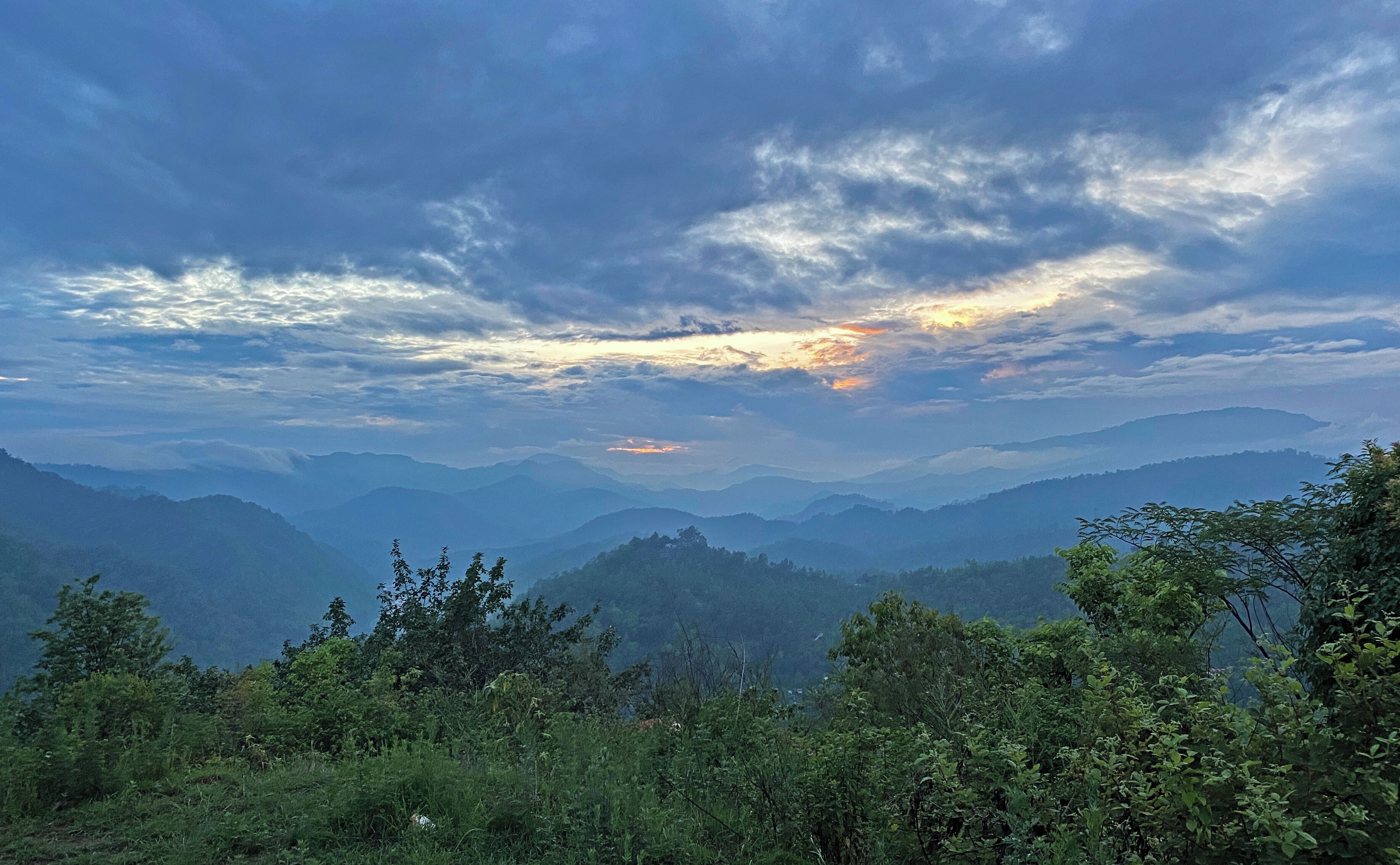 A mountainous landscape with dense forest in the foreground and a cloudy sky above.