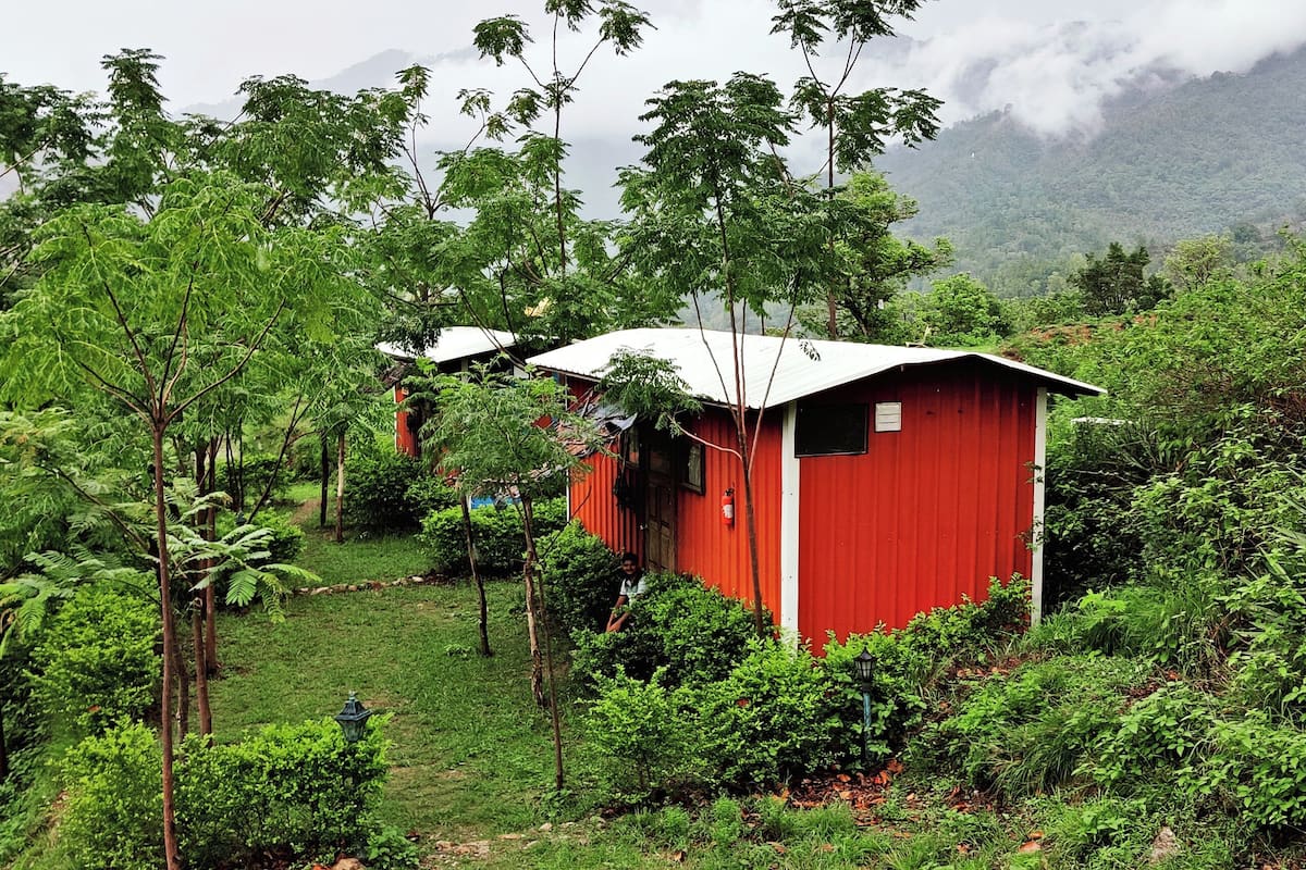 A red cabin surrounded by lush greenery and mountains in the background.