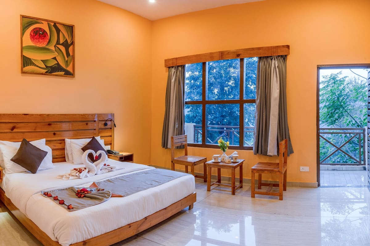 A bedroom with a wooden bed, a wooden headboard, a window with curtains, a small table with a vase, and a view of trees outside.