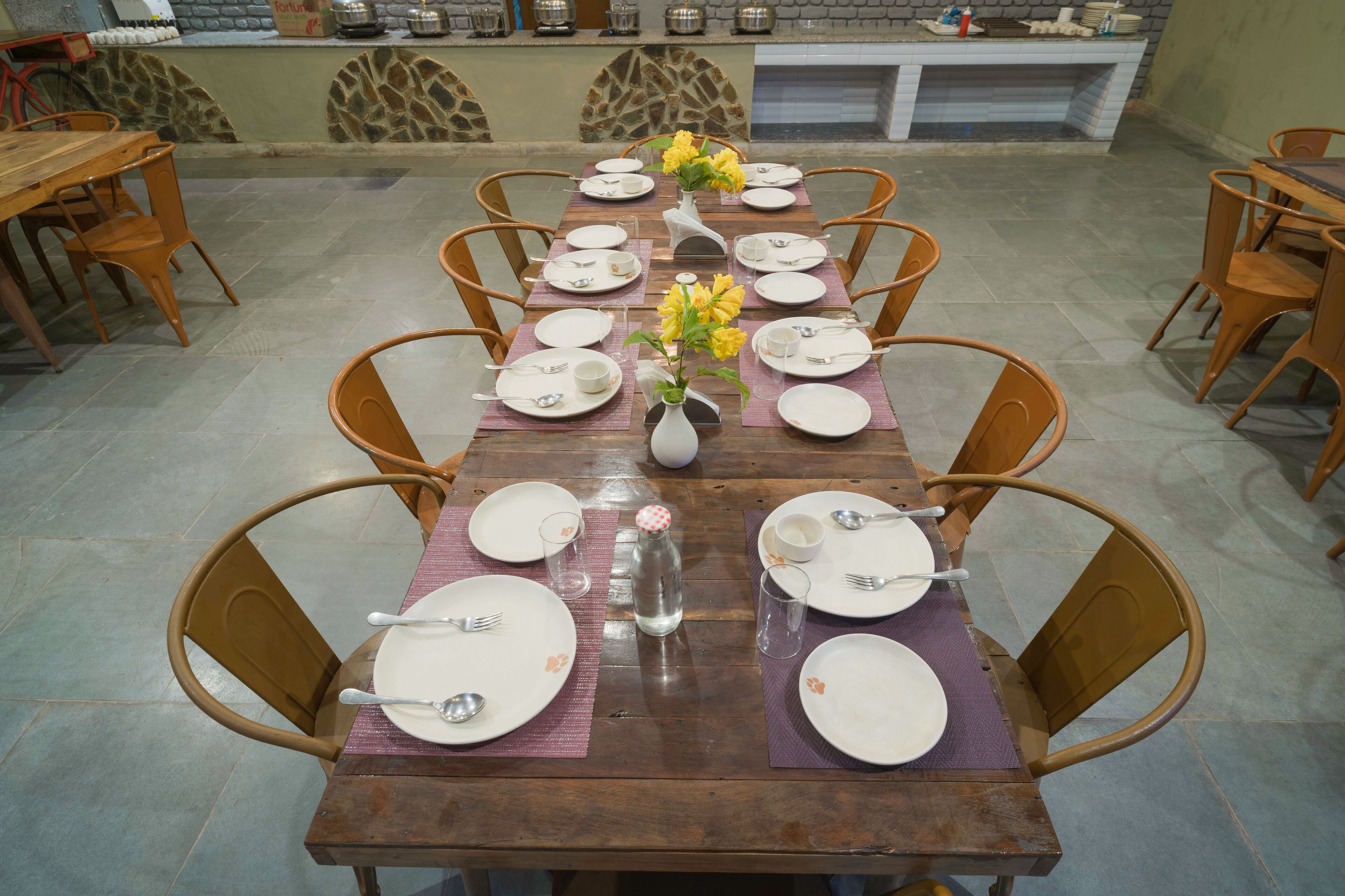 A dining area with a long wooden table set for a meal, surrounded by chairs, with a stone wall and a fireplace in the background.