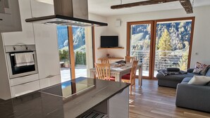 A modern kitchen with a built-in stove, a dining area with a wooden table and chairs, and a living room with a sofa and a large window overlooking a mountainous landscape. - Beautiful chalet - The mountains on the doorstep  (Vintl/Pfunders)