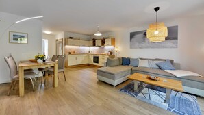 A modern living room with a dining area, a kitchen in the background, and a large window. - Strandküken, Germany (Breege)