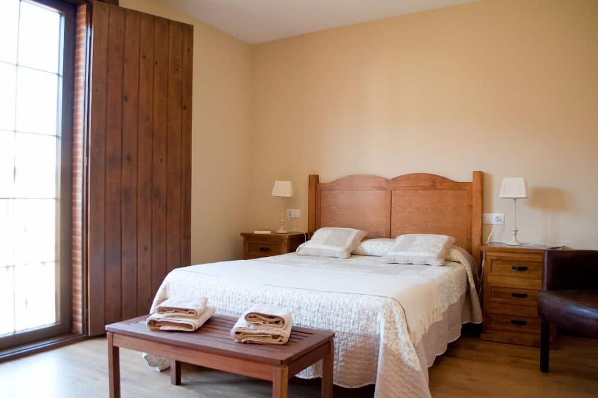 A bedroom with a wooden headboard bed, two bedside tables with lamps, a wooden bench, and a window with blinds.