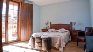 Un dormitorio con una cama de madera, dos mesitas de noche con lámparas, una silla y un ventanal con vistas a edificios.