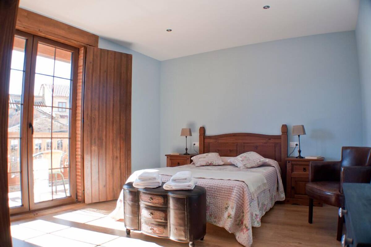 A bedroom with a wooden bed, two bedside tables with lamps, a chair, and a large window with a view of buildings.
