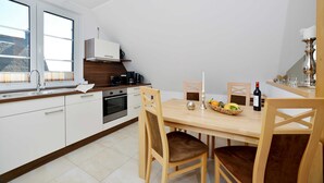 A modern kitchen with white cabinets, a wooden dining table, and a window with blinds. - Boddenbrise Vacation Rental - General Suite (Breege)