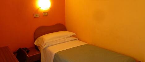 A small, single-bed room with a wooden headboard, a desk with a chair, and a wall-mounted light fixture.