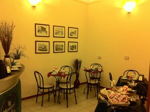 A hotel dining area with tables set for breakfast, framed pictures on the wall, and a coffee station.