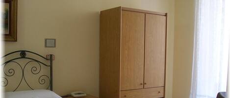 A bedroom with a bed, a wooden wardrobe, a bedside table with a phone, and a window with curtains.