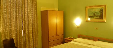 Quarto de hotel com uma cama, guarda-roupa de madeira, cadeira, mesa de cabeceira, abajur e um quadro na parede.