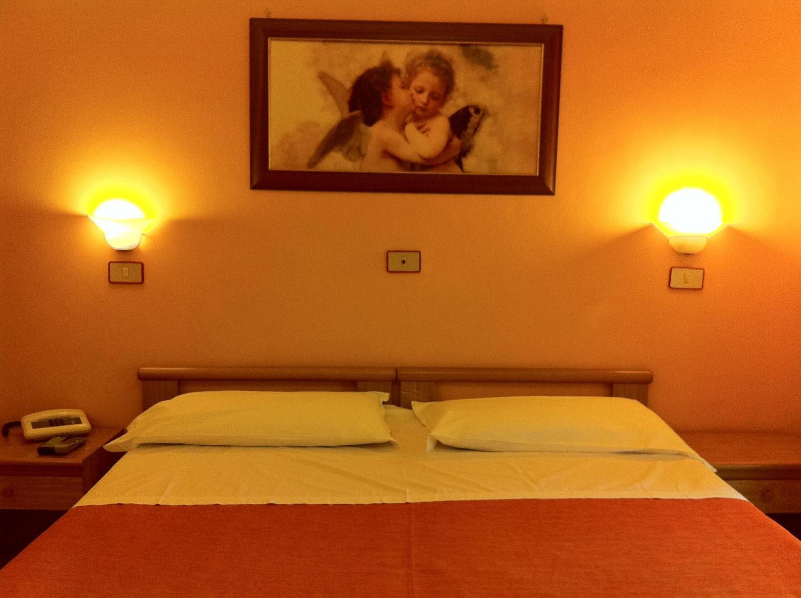 Quarto de hotel com cama, duas luminárias de cabeceira, um quadro na parede e um telefone na mesa de apoio.