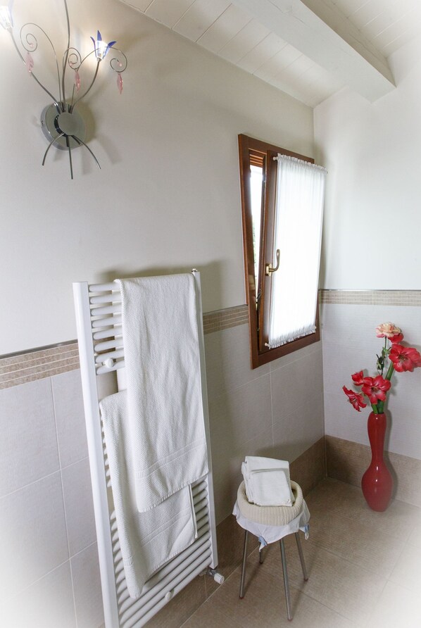 A bathroom with a white towel rack, a small stool with a towel, a red vase with flowers, and a wall-mounted light fixture.