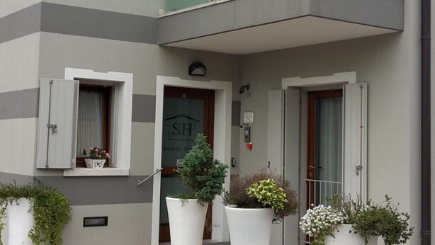 A modern apartment building with a grey and white facade, large potted plants, and a small balcony. - Sweet Home (Treviso)