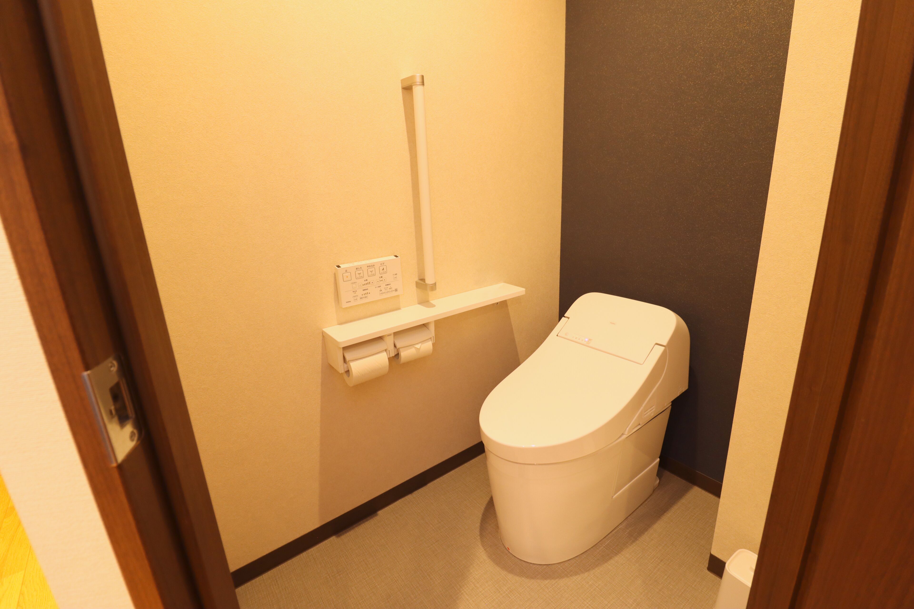 A small, compact bathroom with a toilet, a shelf with toilet paper, and a wall-mounted toilet paper holder.