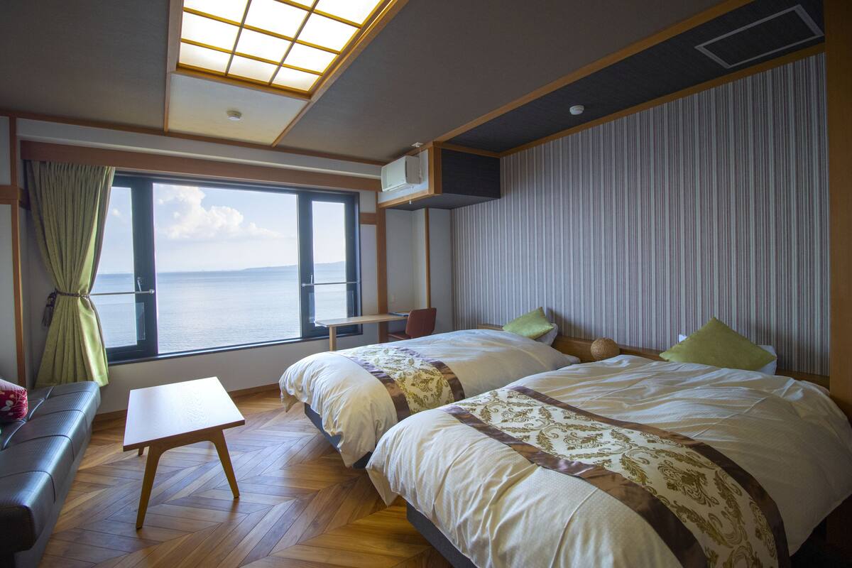 Una habitación con dos camas, una mesita, un sofá, un ventanal con vista al mar y un tragaluz.