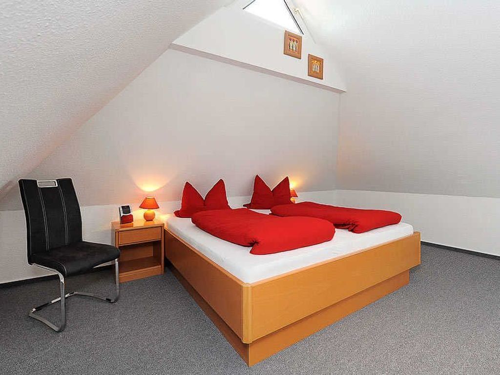 A modern bedroom with a wooden bed, red pillows, a black chair, a nightstand with a lamp, and a small table.
