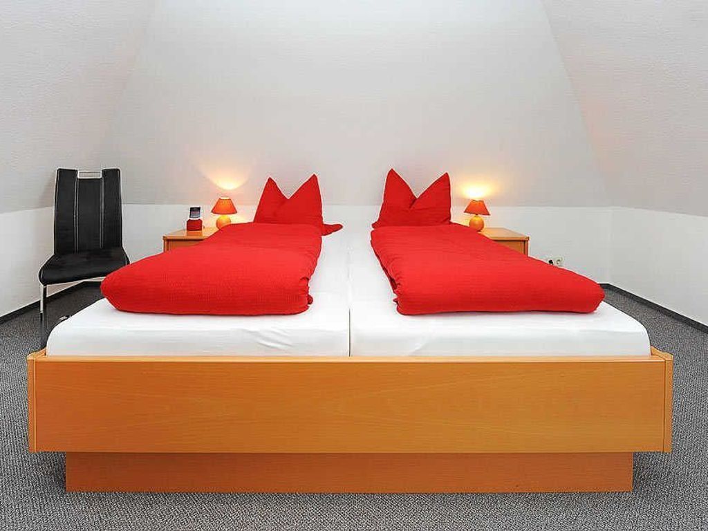 A double bed with red bedding, two bedside tables with lamps, a black chair, and a white wall.