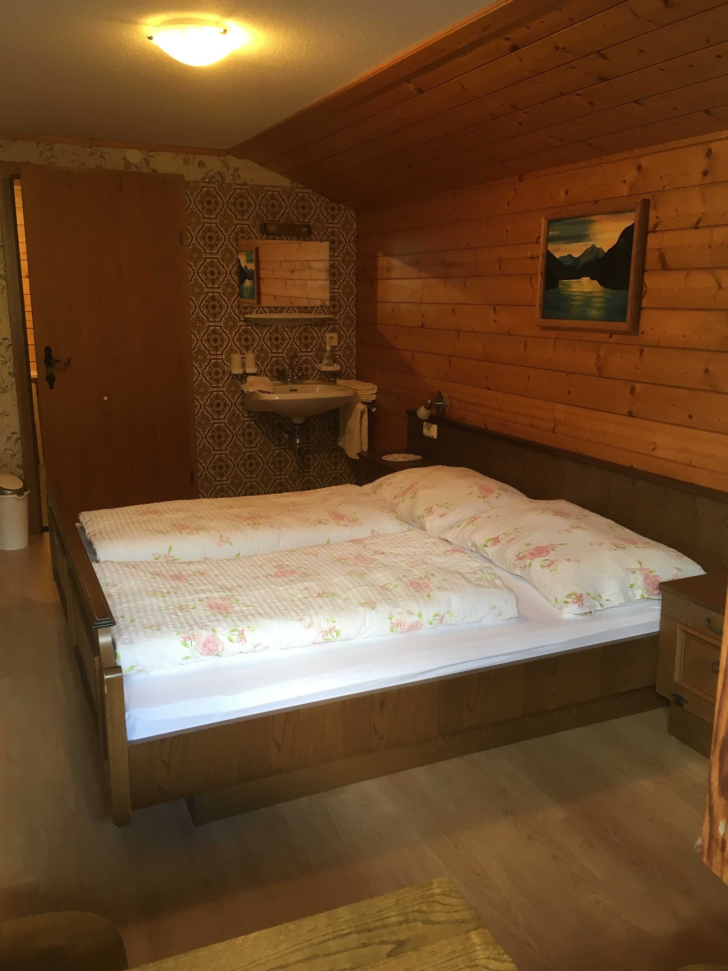 A wooden cabin bedroom with a bed, a wooden dresser, a bathroom with a sink, and a painting on the wall.