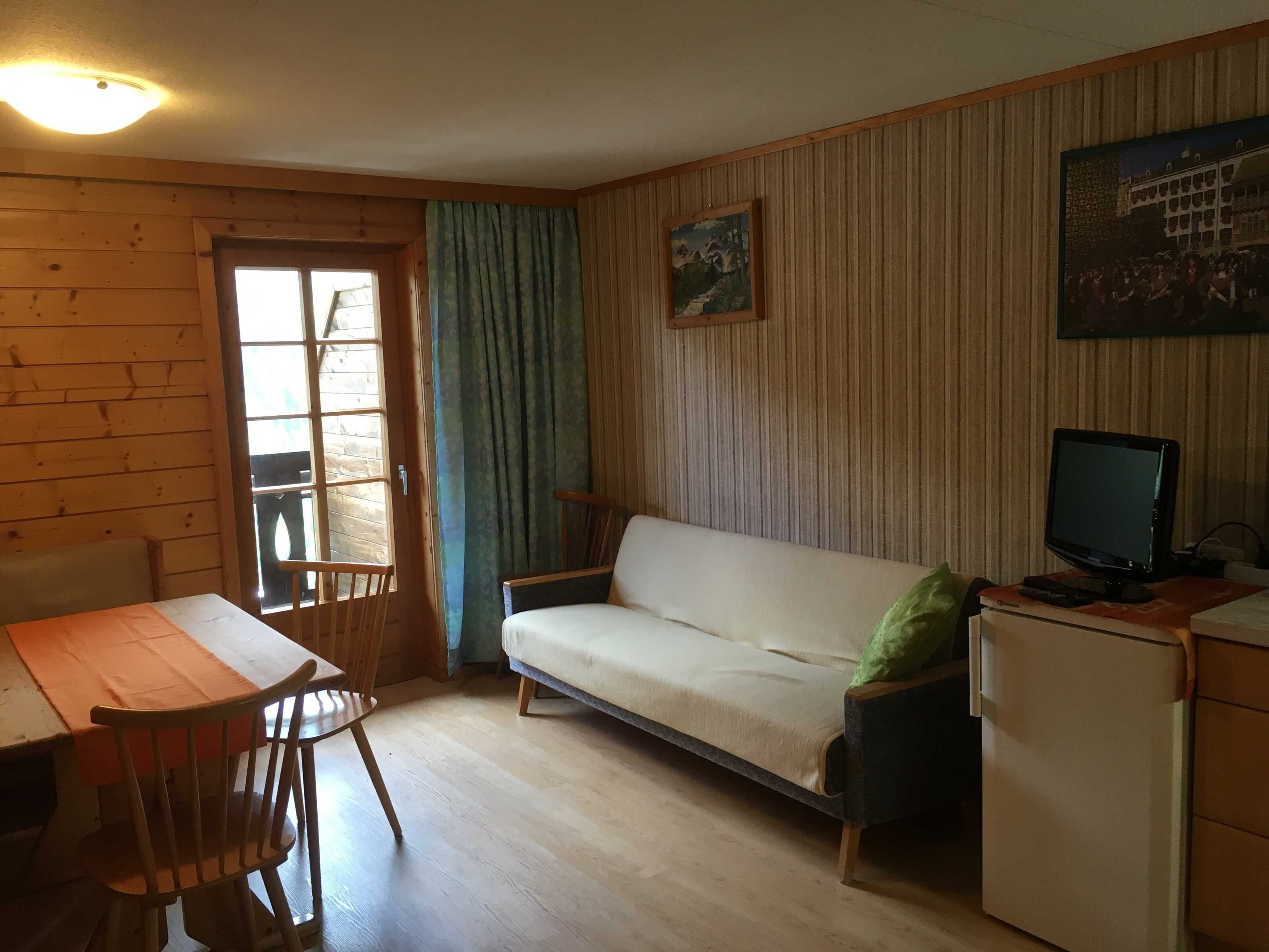 Une pièce avec un plancher en bois, un canapé blanc, une table à manger avec des chaises, un téléviseur posé sur un meuble et un réfrigérateur.
