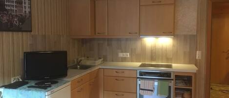 Cocina compacta con armarios de madera, horno empotrado y un televisor sobre una mesita.