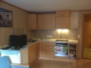 Una cuina petita amb armaris de fusta, un forn integrat i una televisió sobre una taula.