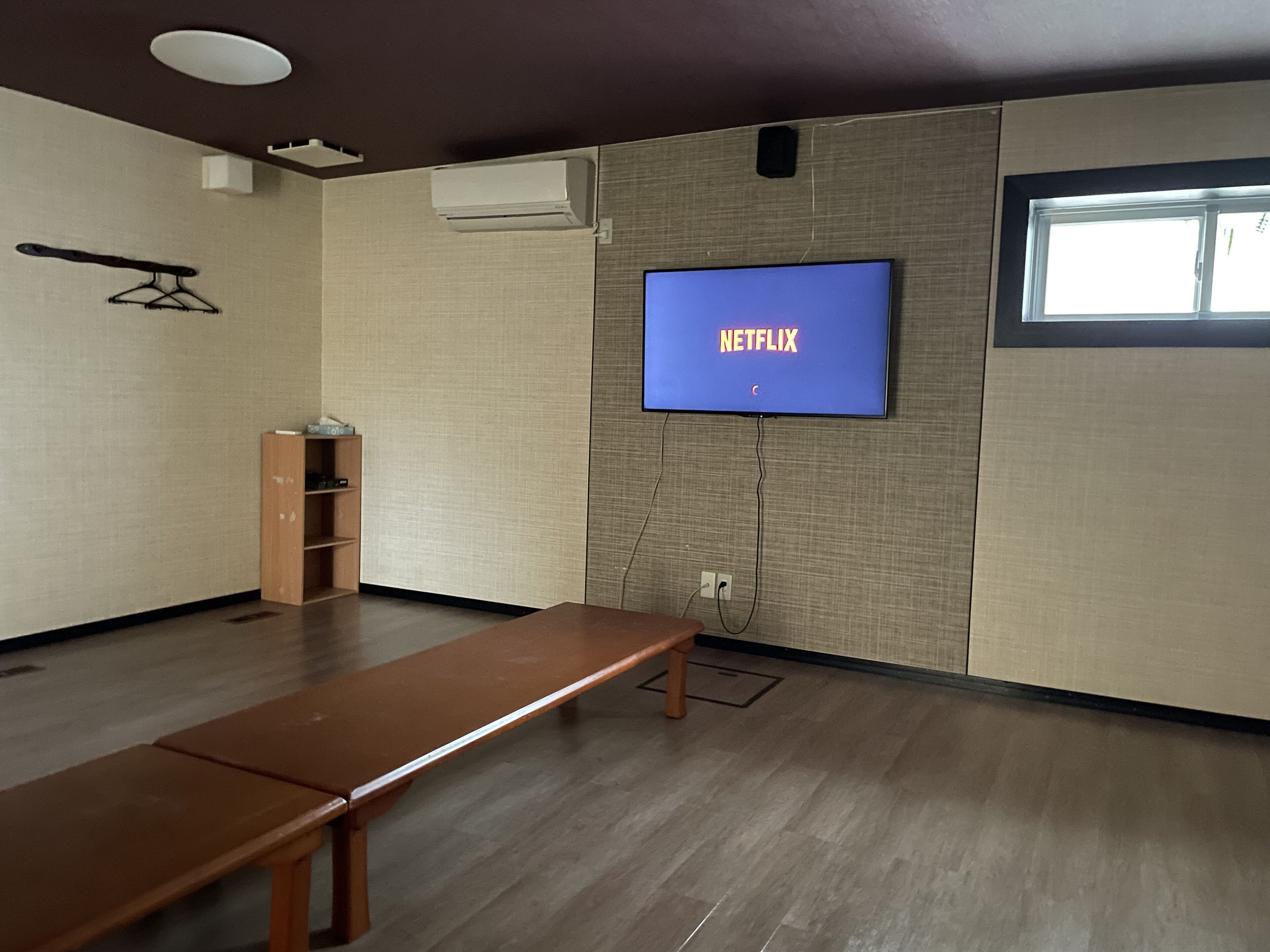 Een kamer met een houten bankje, een televisie waarop 'NETFLIX' staat, een wandairconditioner en een raam.