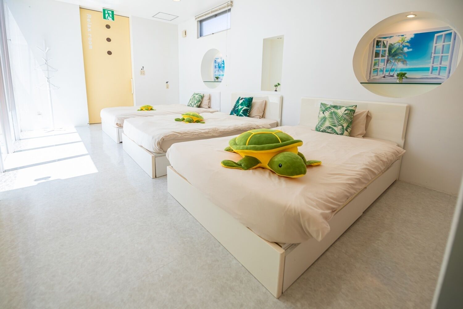 A hotel room with two beds, each with a green turtle plush toy, a window with a view of palm trees, and a circular wall art depicting a beach scene.
