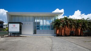 Un edificio moderno con un amplio muro de vidrio, un letrero con caracteres japoneses y una palmera en primer plano.