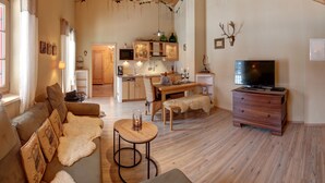 Ein gemütliches Wohnzimmer mit Holzbalkendecke, einem Fernseher, einer Couch und einem Essbereich mit einer Küche im Hintergrund.
