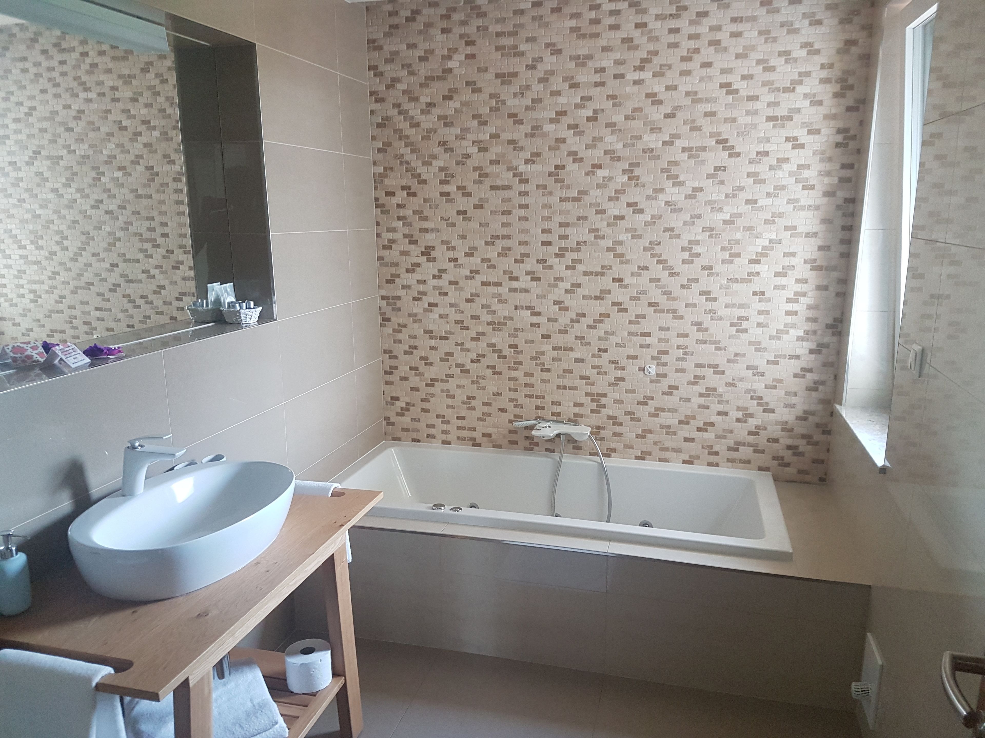 A bathroom with a large white bathtub, a wooden vanity with a white sink, and a mirror above the sink. The walls are tiled with a patterned design.