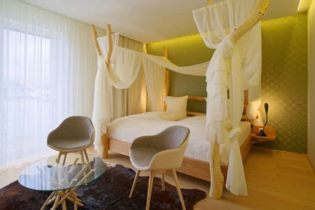 Une chambre à coucher avec un lit à baldaquin, deux fauteuils, une table en verre et une vue sur l’extérieur.