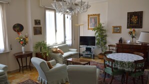 Ein Wohnzimmer mit einer Couch, Sesseln, einem Fernseher, einer Chandelier und einem Esstisch mit Stühlen.