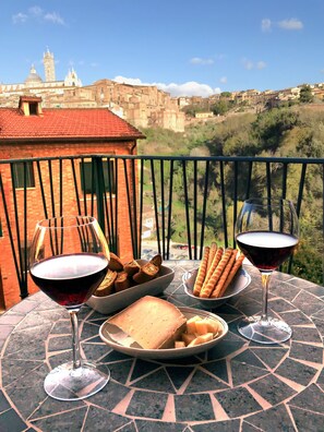 Une table avec des verres à vin, du pain et un bol de collations, placée sur un balcon donnant sur un paysage urbain avec des bâtiments historiques.