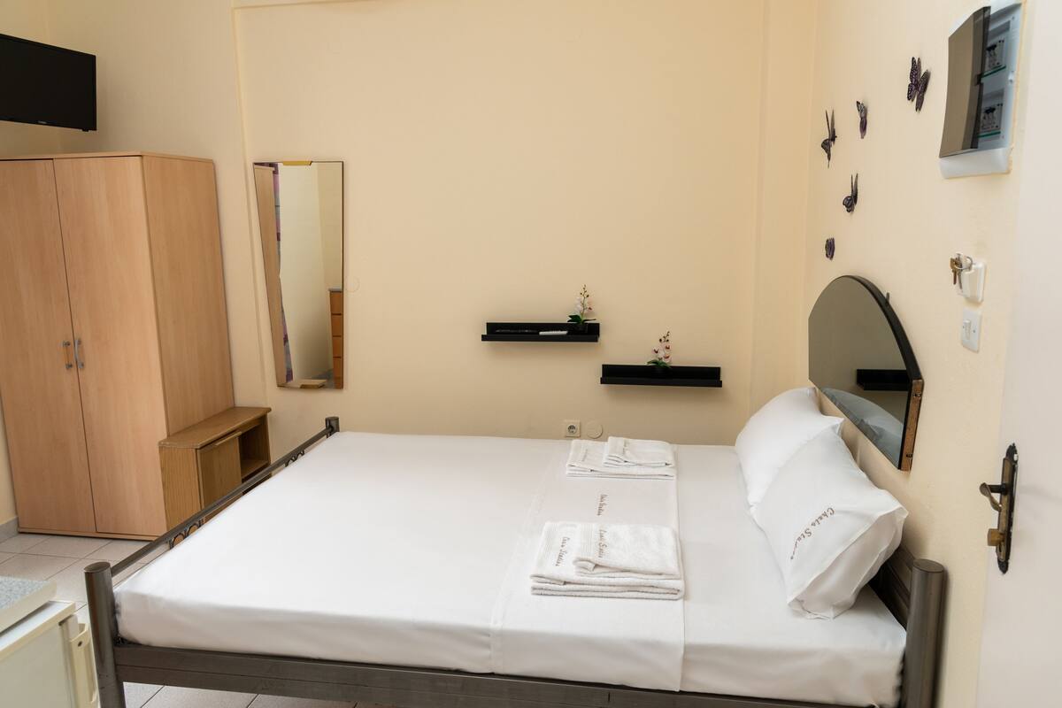 A neatly made bed with white linens and towels, a wooden wardrobe, a small shelf with a plant, a mirror, and butterfly wall decor.
