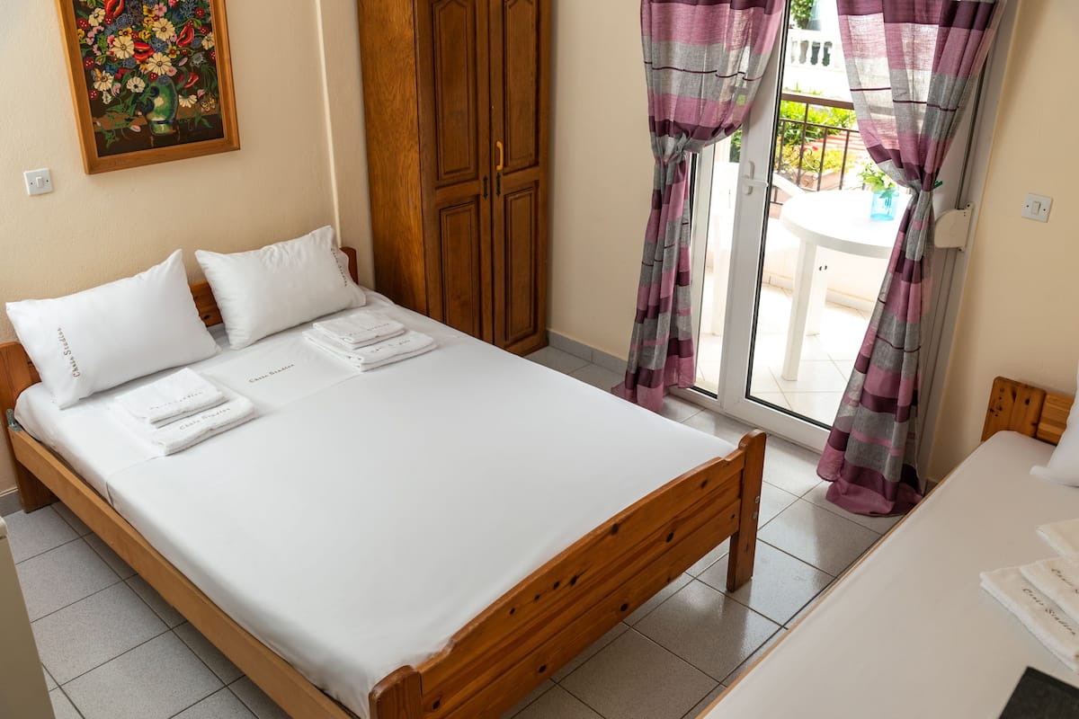 A neatly made bed with white linens and pillows, a wooden wardrobe, and a view through a glass door to an outdoor area.