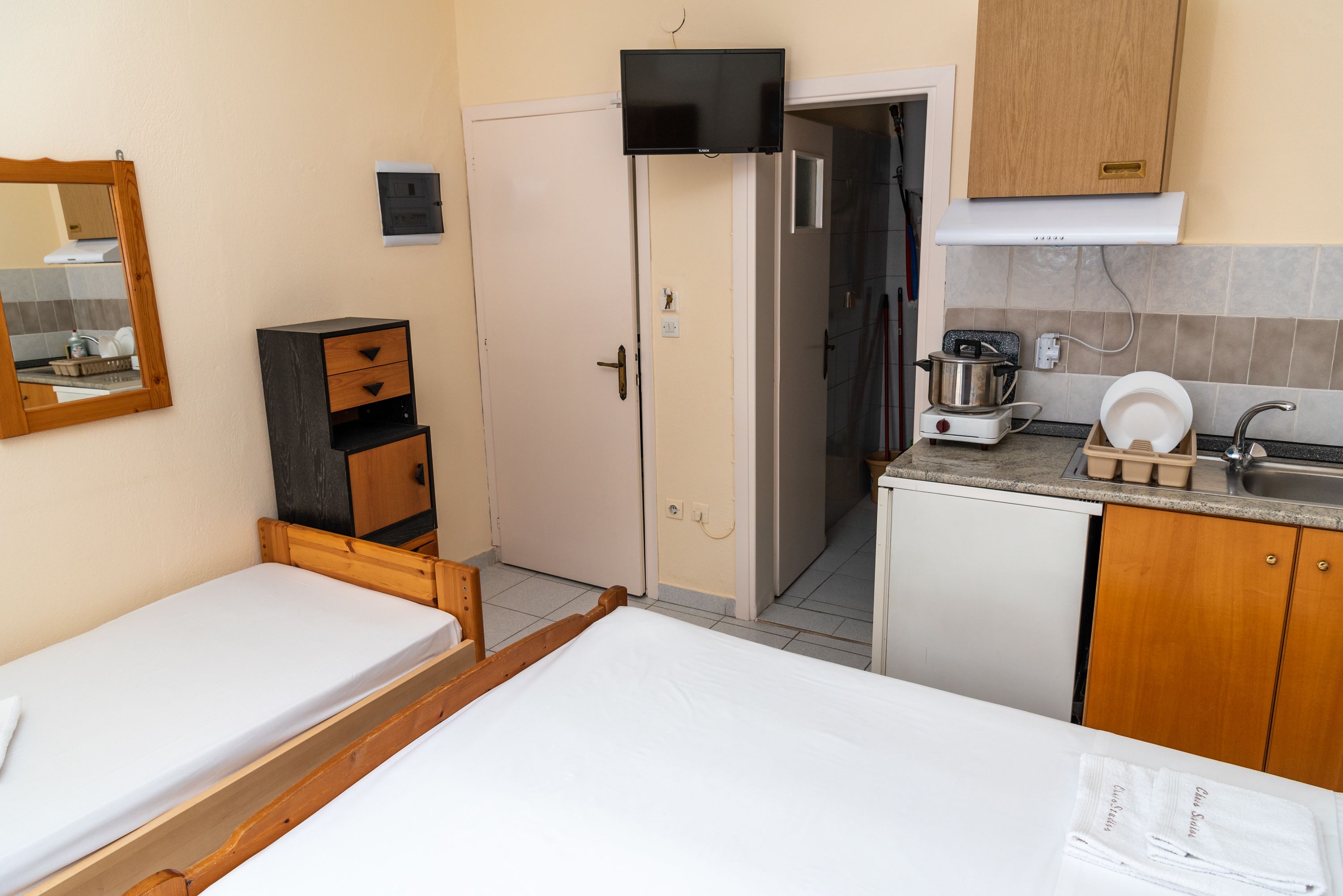 A compact hotel room with a single bed, a small wooden dresser, a kitchenette with a stove and sink, and a door leading to a bathroom.