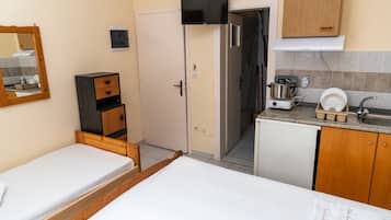 A compact hotel room with a single bed, a small wooden dresser, a kitchenette with a stove and sink, and a door leading to a bathroom.