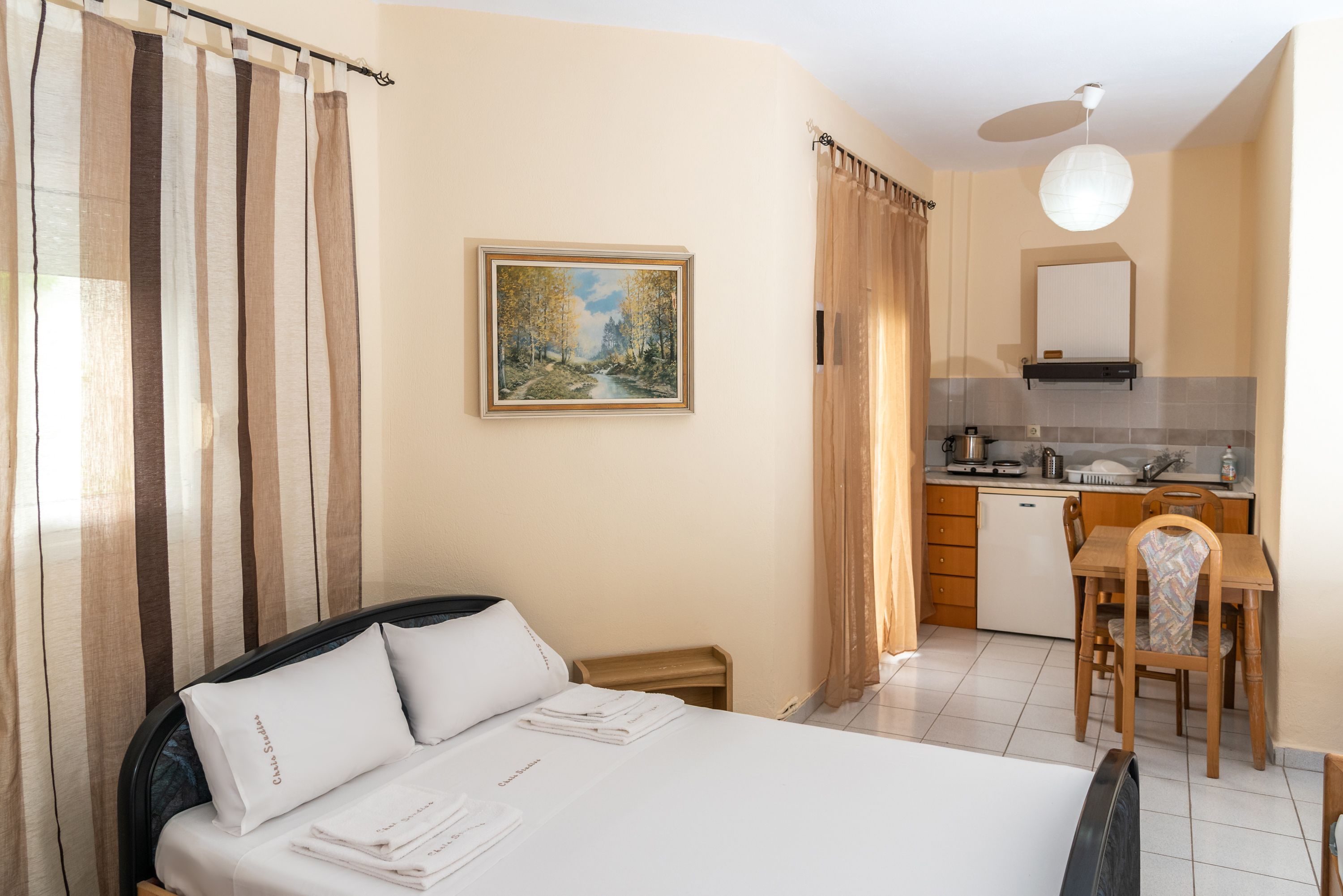 A hotel room with a bed, a desk, a chair, a painting on the wall, and a kitchen area.