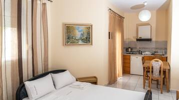 A hotel room with a bed, a desk, a chair, a painting on the wall, and a kitchen area.