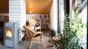 Ett mysigt rum med ett träpanelat tak, en öppen spis, en fåtölj i rotting, en bokhylla och ett stort fönster med utsikt.