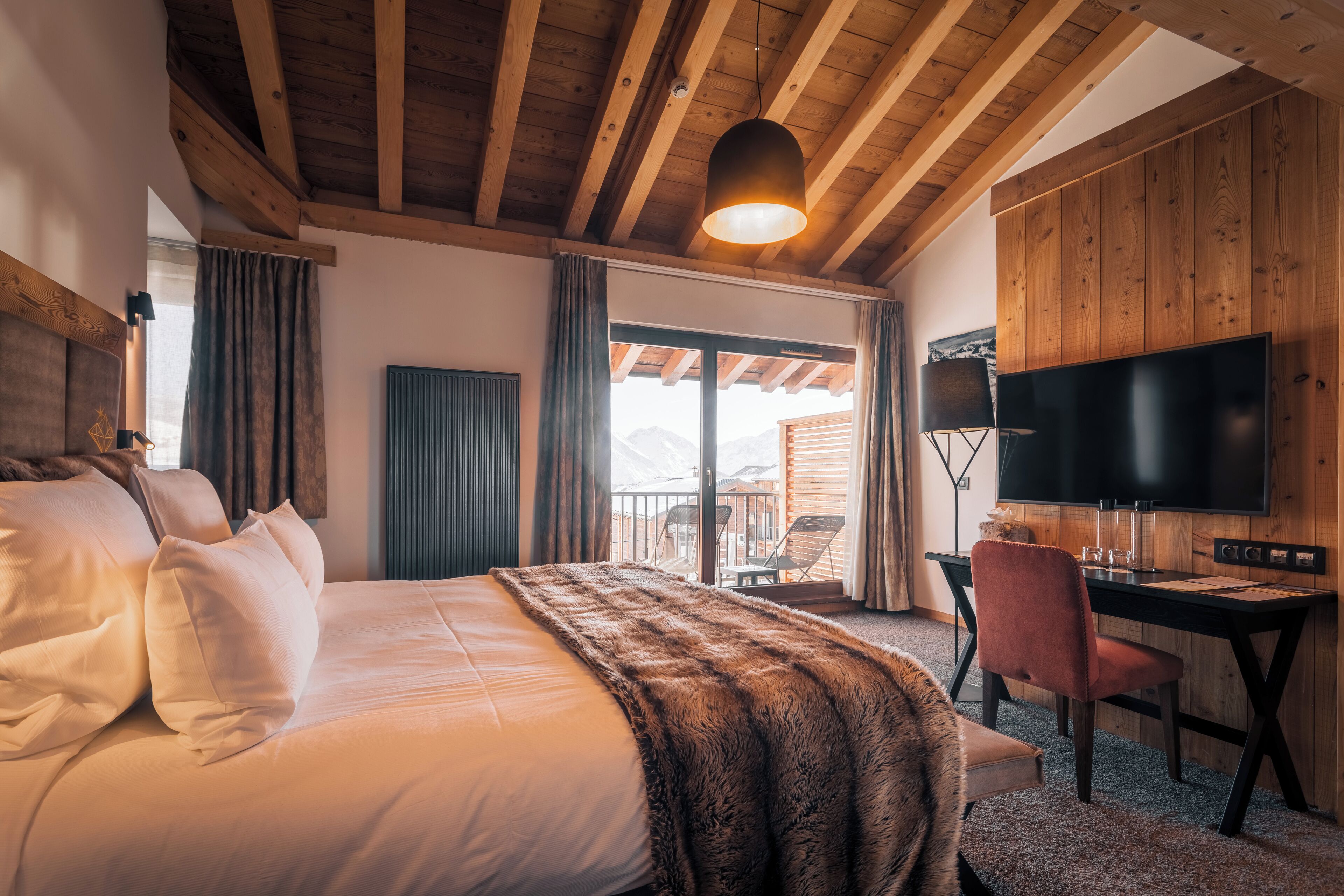 Une chambre avec un grand lit, un bureau, une télévision et un balcon offrant une vue sur les montagnes.