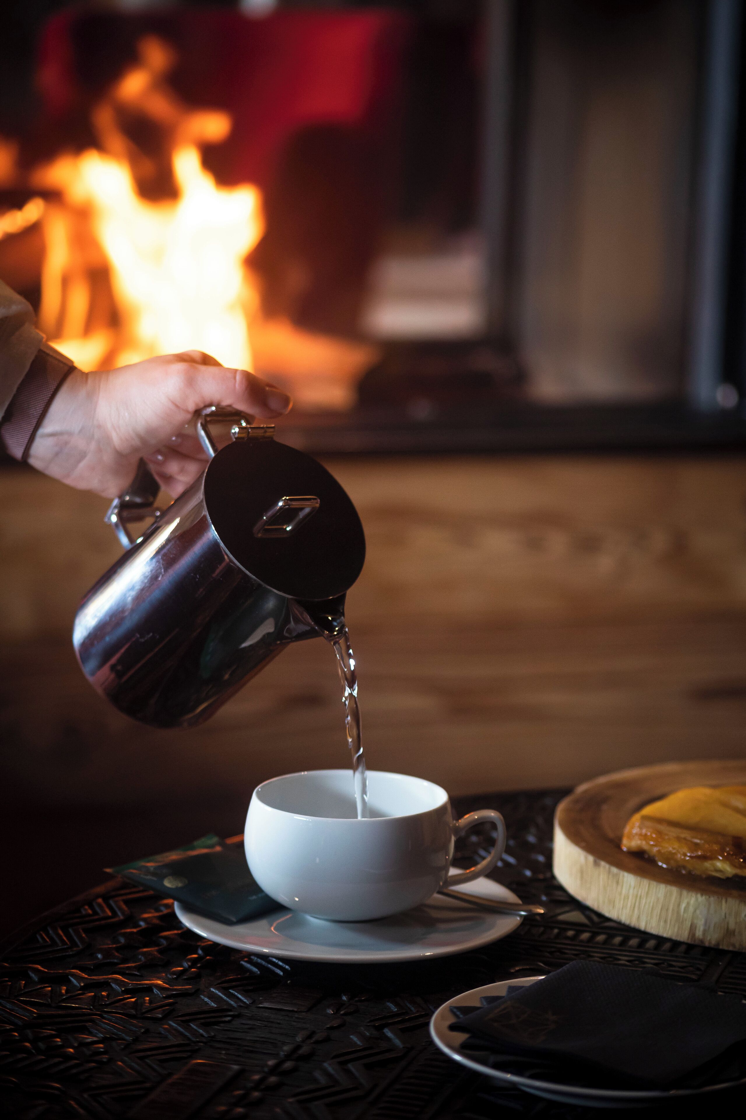 Une main verse un liquide d’un pichet en métal dans une tasse blanche posée sur une table, à côté d’une assiette et d’une serviette.
