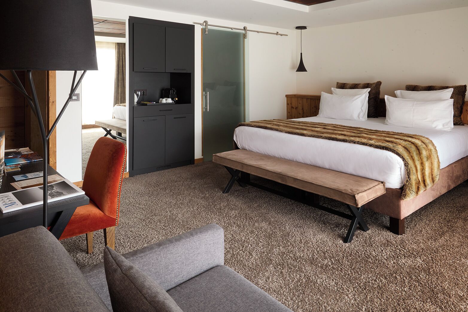 Quarto de hotel com cama, escrivaninha com cadeira, um banco, guarda-roupa e luminária suspensa.