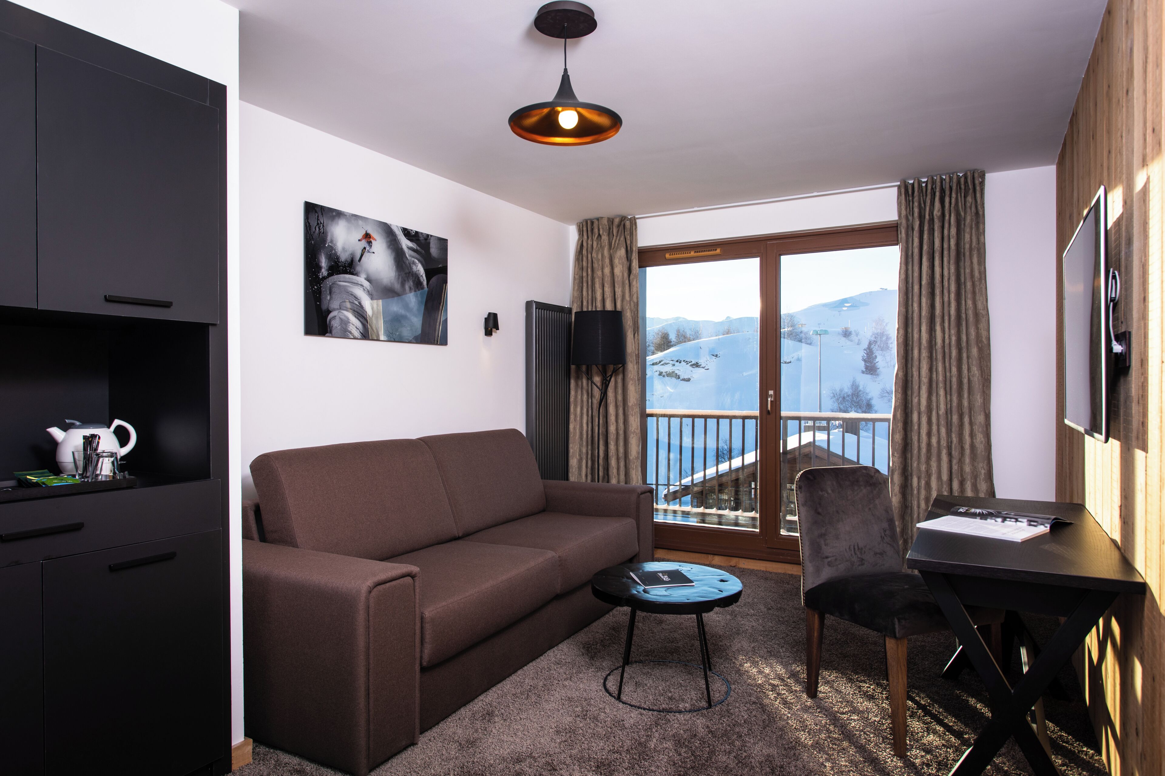 Un salon moderne avec un canapé, une petite table, un téléviseur et un balcon offrant une vue sur un paysage enneigé.