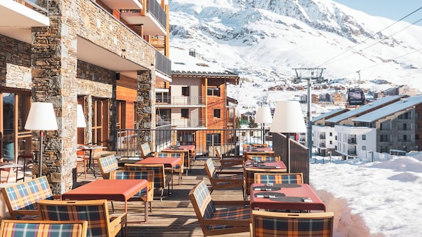 A snow-covered mountain with a ski lift in the background, a wooden deck with outdoor seating, and a stone building with balconies. - Hôtel Daria-I Nor by Les Etincelles (Huez)